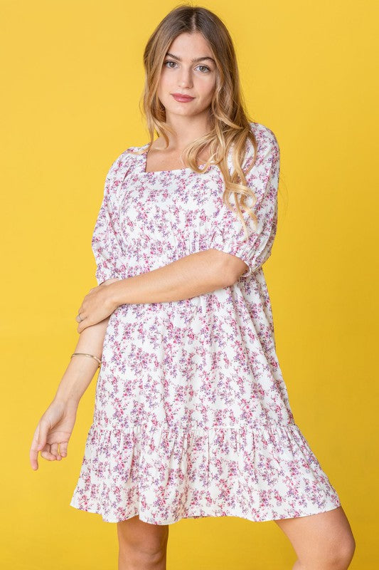 A person in a Ditsy Floral Squared Neck Mini Dress stands against a brick wall adorned with green foliage, gently holding strands of their hair.