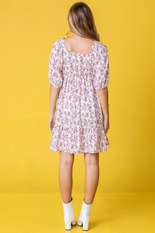 A person in a Ditsy Floral Squared Neck Mini Dress stands against a brick wall adorned with green foliage, gently holding strands of their hair.