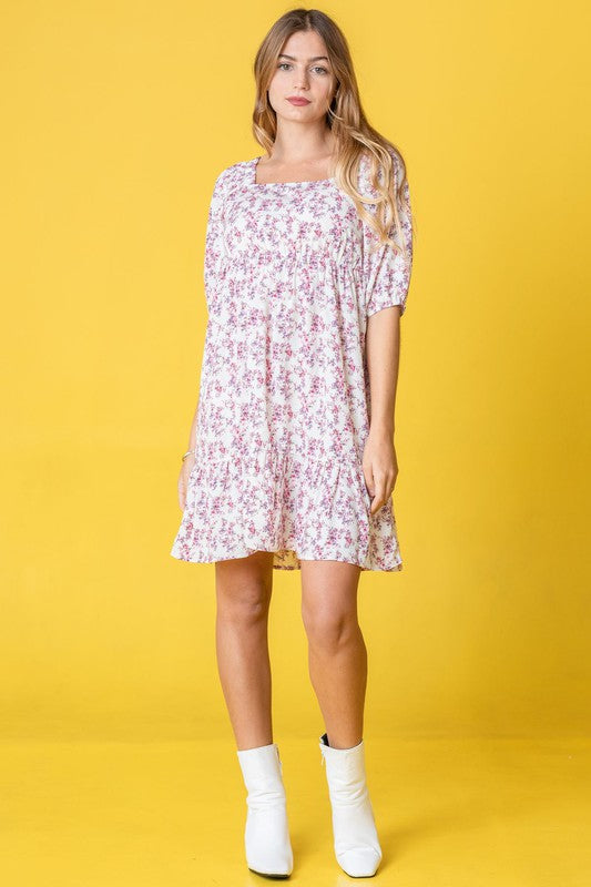 A person in a Ditsy Floral Squared Neck Mini Dress stands against a brick wall adorned with green foliage, gently holding strands of their hair.