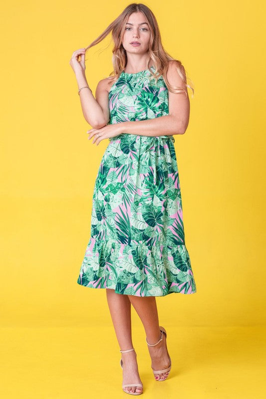 A woman wearing the Plus Halter Neck Tropical Sash Dress in a lively green and pink leaf pattern poses against a sunny yellow background, gently holding a strand of her hair.