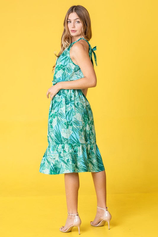 A person wearing a Halter Neck Tropical Sash Dress featuring a green leafy pattern stands against a yellow background.