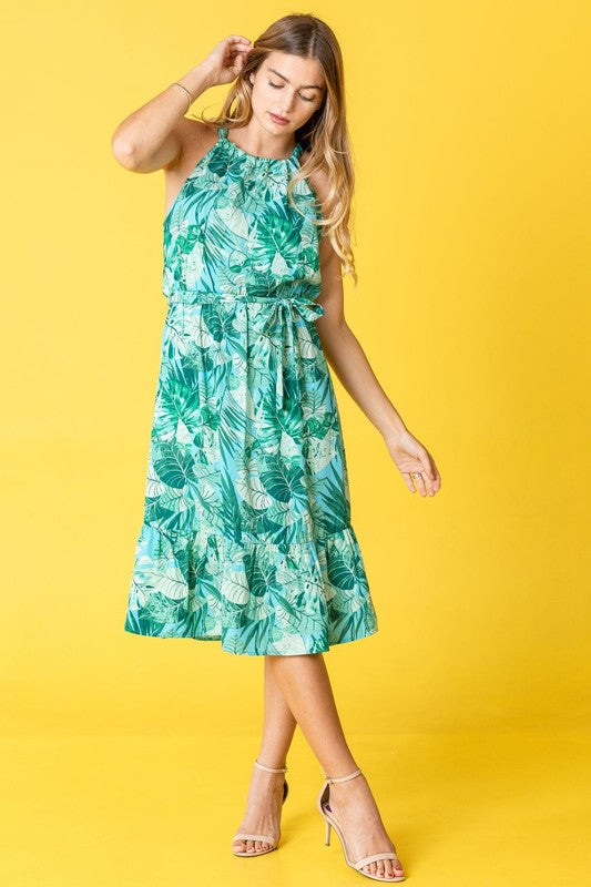 A woman wearing the Plus Halter Neck Tropical Sash Dress in a lively green and pink leaf pattern poses against a sunny yellow background, gently holding a strand of her hair.