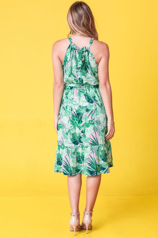 A woman wearing the Plus Halter Neck Tropical Sash Dress in a lively green and pink leaf pattern poses against a sunny yellow background, gently holding a strand of her hair.