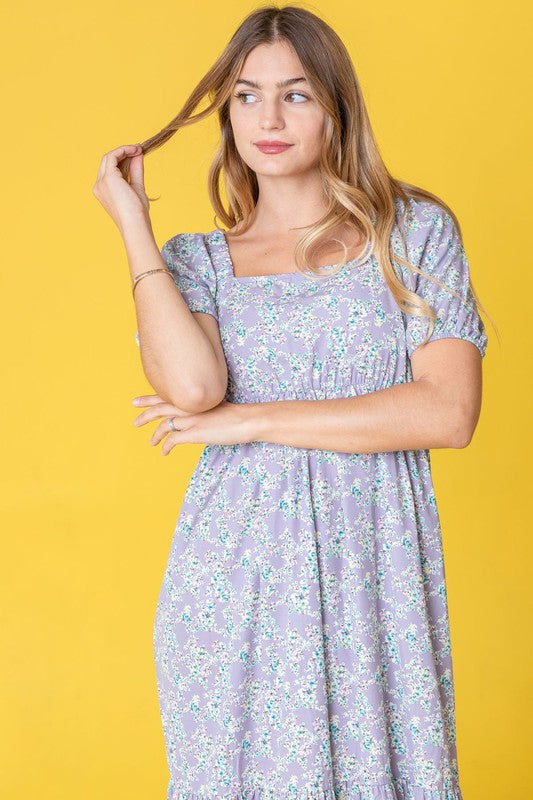 A woman in a Ditsy Floral Square Neck Sundress, crafted from 100% polyester, stands against a yellow background, highlighting its elegant light blue floral design.