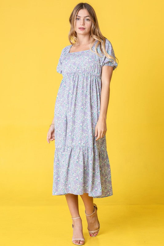 A woman in a Ditsy Floral Square Neck Sundress, crafted from 100% polyester, stands against a yellow background, highlighting its elegant light blue floral design.