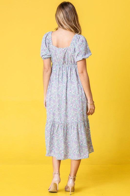 A woman in a Ditsy Floral Square Neck Sundress, crafted from 100% polyester, stands against a yellow background, highlighting its elegant light blue floral design.