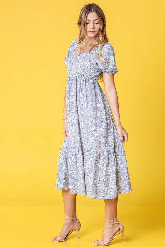 A woman in a Ditsy Floral Square Neck Sundress, crafted from 100% polyester, stands against a yellow background, highlighting its elegant light blue floral design.