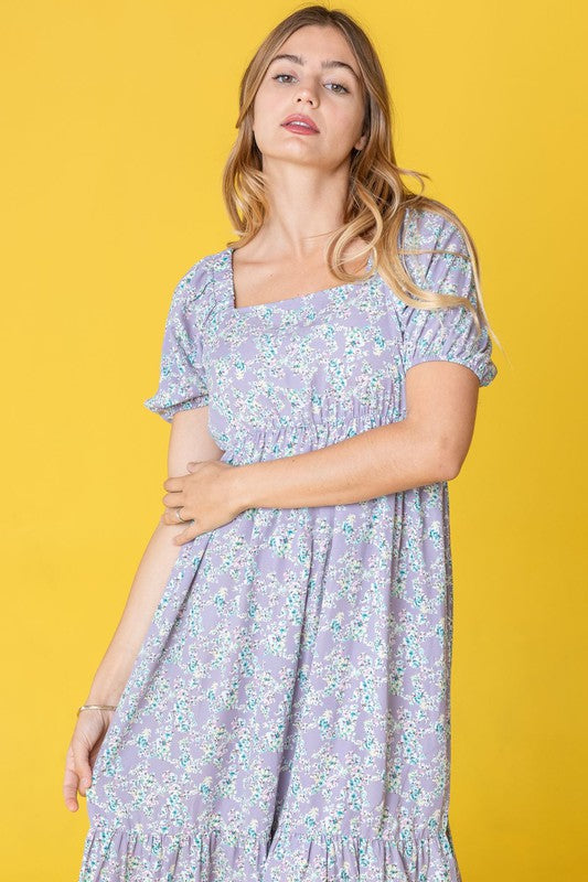 A woman in a Ditsy Floral Square Neck Sundress, crafted from 100% polyester, stands against a yellow background, highlighting its elegant light blue floral design.