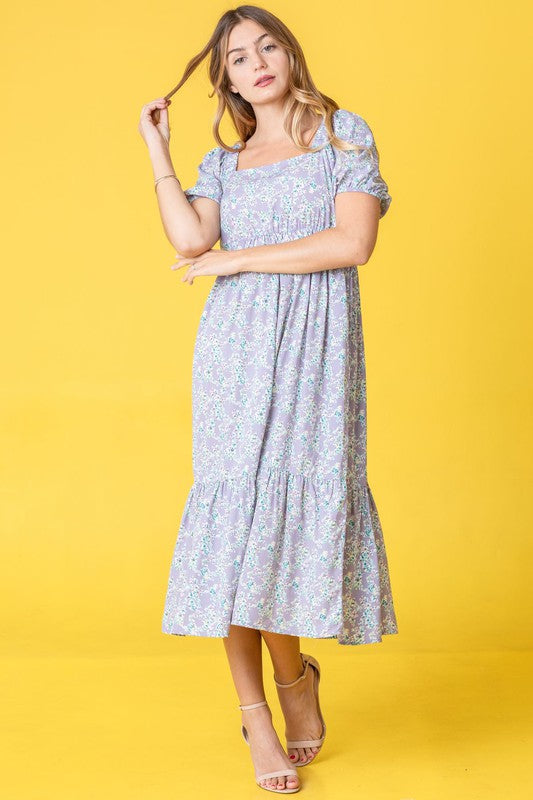 A woman in a Ditsy Floral Square Neck Sundress, crafted from 100% polyester, stands against a yellow background, highlighting its elegant light blue floral design.