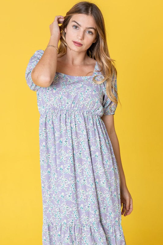 A woman in a Ditsy Floral Square Neck Sundress, crafted from 100% polyester, stands against a yellow background, highlighting its elegant light blue floral design.