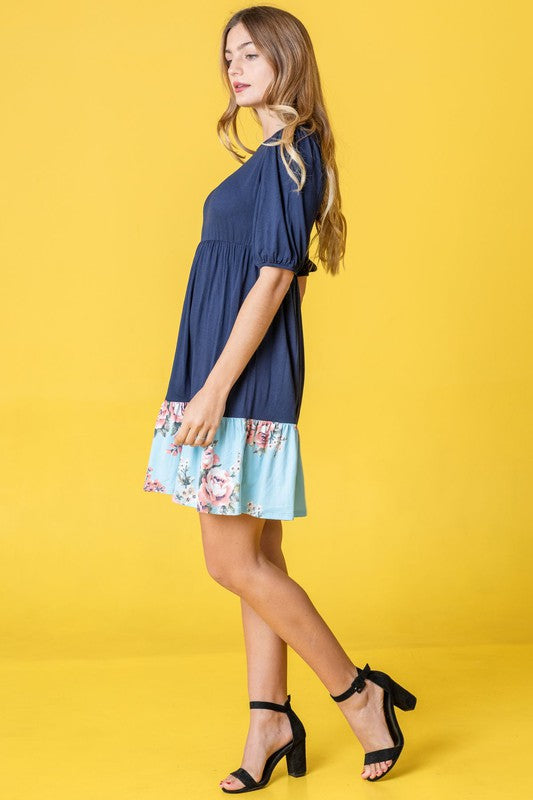 A person wearing the Floral Two Tone Tiered Mini Dress, featuring puff sleeves and a vibrant tie-dye hem, stands against a beige background, smiling.