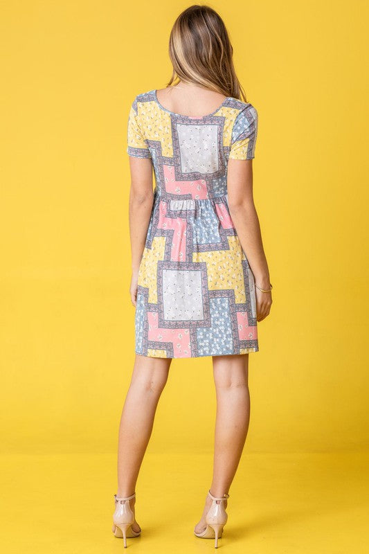 A woman wearing a Plus Short Sleeve Floral Patch Work Mini Dress stands against a yellow background, holding a strand of her hair and looking to the side.