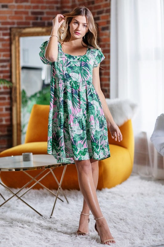 A woman poses against a pink background, dressed in the Plus Tropical Flutter Sleeve Tiered Midi Dress in a vibrant green print and paired with black sandals.