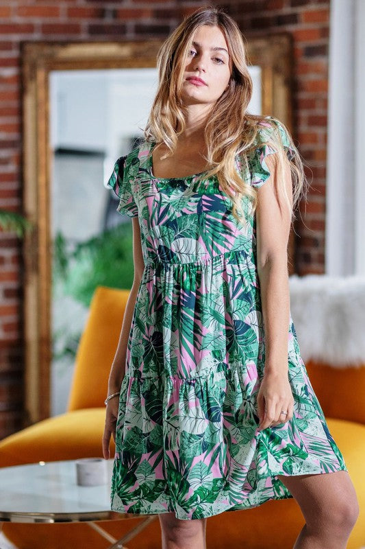 A person with long hair stands wearing a Tropical Flutter Sleeve Tiered Midi Dress in a green and pink leaf pattern. Behind them is an orange chair and a brick wall featuring a framed mirror, reflecting their vivid style.