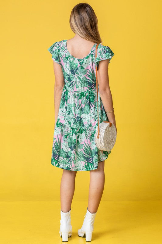 A woman poses against a pink background, dressed in the Plus Tropical Flutter Sleeve Tiered Midi Dress in a vibrant green print and paired with black sandals.