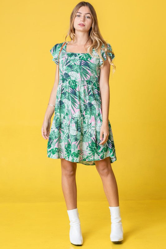 A person with long hair stands wearing a Tropical Flutter Sleeve Tiered Midi Dress in a green and pink leaf pattern. Behind them is an orange chair and a brick wall featuring a framed mirror, reflecting their vivid style.