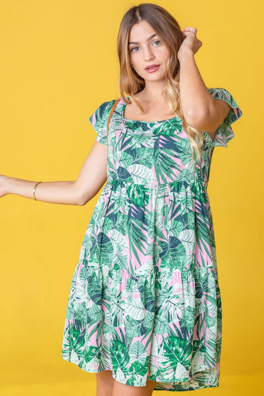 A person with long hair stands wearing a Tropical Flutter Sleeve Tiered Midi Dress in a green and pink leaf pattern. Behind them is an orange chair and a brick wall featuring a framed mirror, reflecting their vivid style.