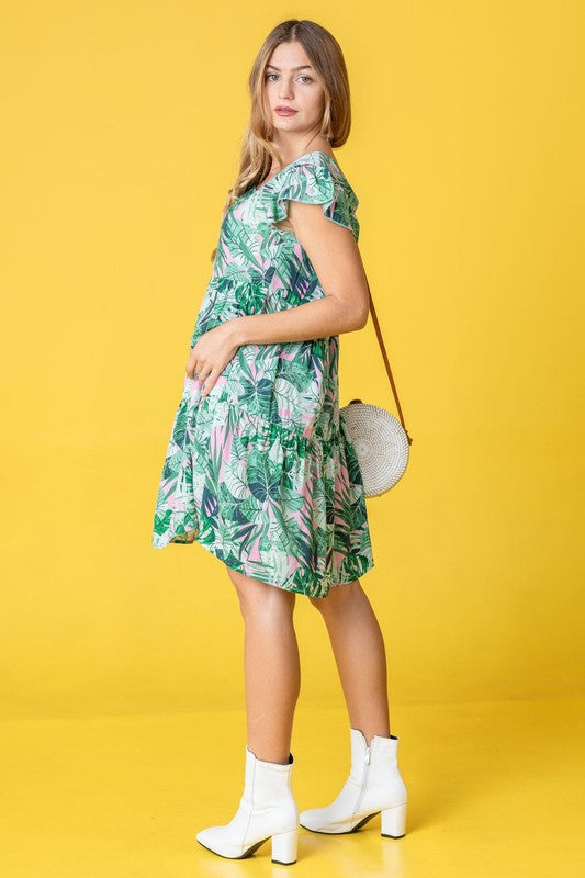 A person with long hair stands wearing a Tropical Flutter Sleeve Tiered Midi Dress in a green and pink leaf pattern. Behind them is an orange chair and a brick wall featuring a framed mirror, reflecting their vivid style.