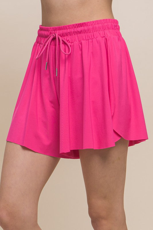 A young woman in a yellow top and Activewear Two In One Drawstring Shorts holding a tennis racket, smiling at the camera.