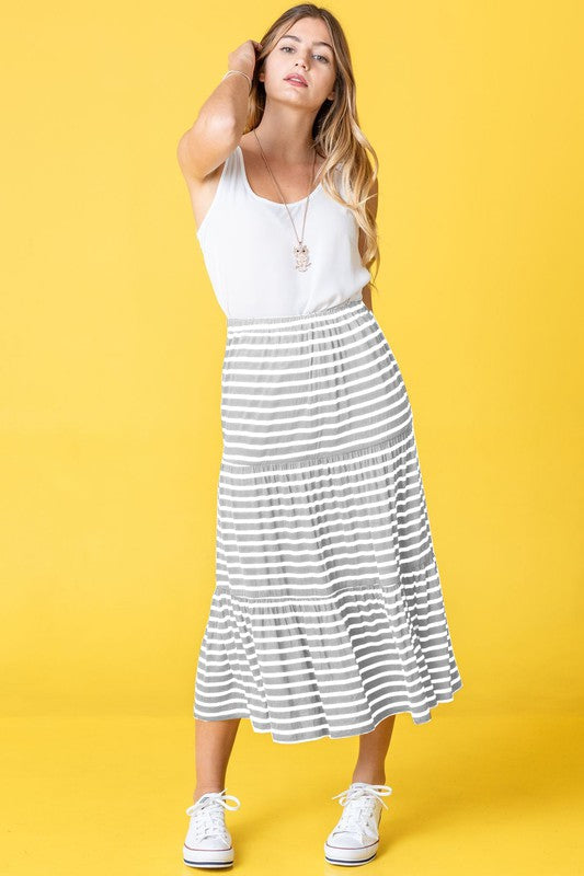 A woman poses against a pink backdrop, dressed in a white sleeveless top, the Stripe Maxi Skirt crafted in the USA, and white sneakers.