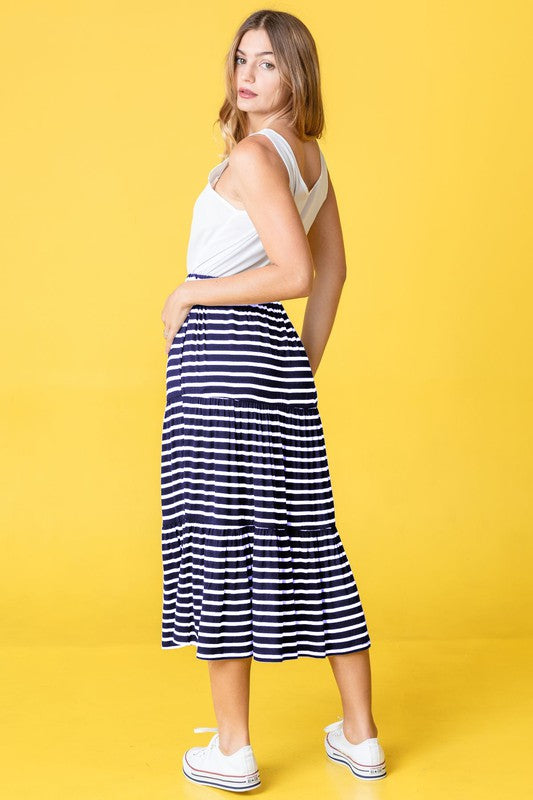 A woman poses against a pink backdrop, dressed in a white sleeveless top, the Stripe Maxi Skirt crafted in the USA, and white sneakers.