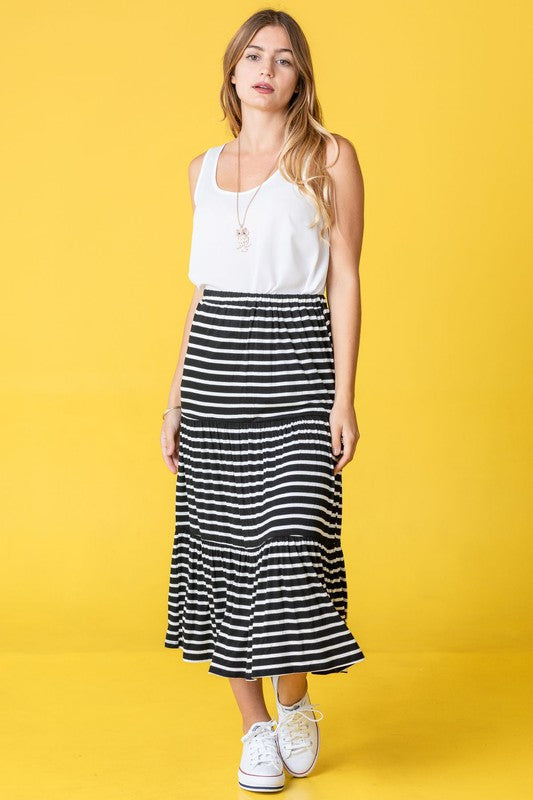 A woman in a white tank top and a Stripe Maxi Skirt stands confidently against a yellow background, accessorized with stylish white sneakers. Made in the United States, her outfit effortlessly combines comfort and elegance.