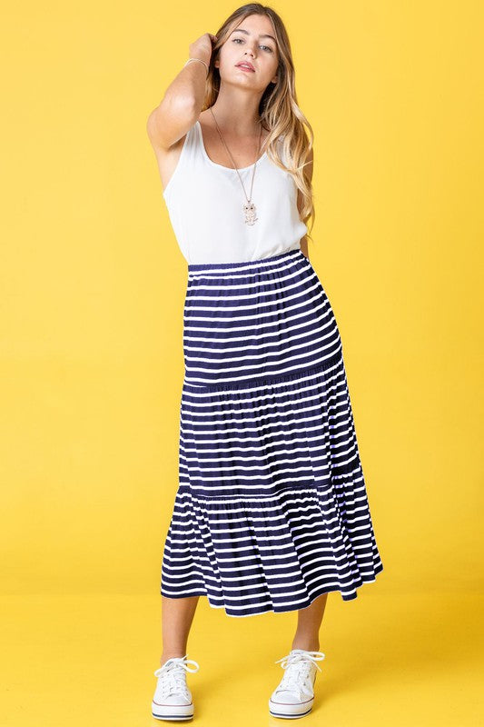 A woman poses against a pink backdrop, dressed in a white sleeveless top, the Stripe Maxi Skirt crafted in the USA, and white sneakers.