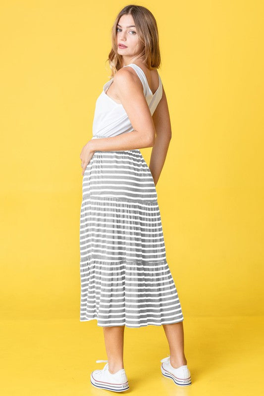 A woman poses against a pink backdrop, dressed in a white sleeveless top, the Stripe Maxi Skirt crafted in the USA, and white sneakers.