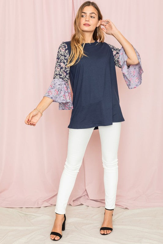 A woman in a Color Block Ruffle Sleeve Tunic, featuring elegant floral bell sleeves, stands against a pink backdrop, gracefully touching her hair.