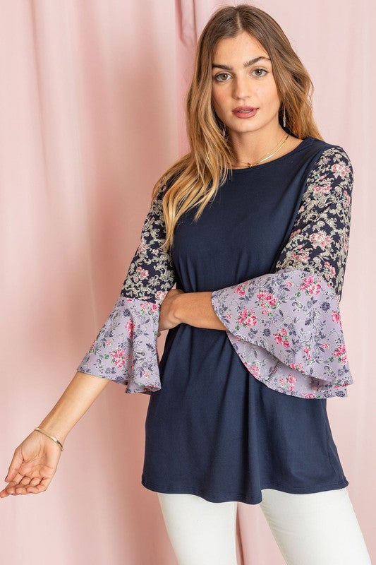 A woman in a Color Block Ruffle Sleeve Tunic, featuring elegant floral bell sleeves, stands against a pink backdrop, gracefully touching her hair.