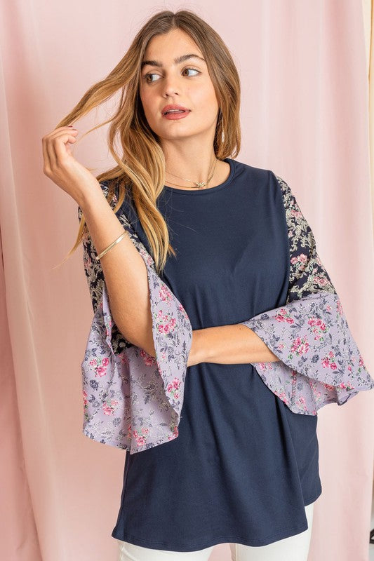 A woman in a Color Block Ruffle Sleeve Tunic, featuring elegant floral bell sleeves, stands against a pink backdrop, gracefully touching her hair.