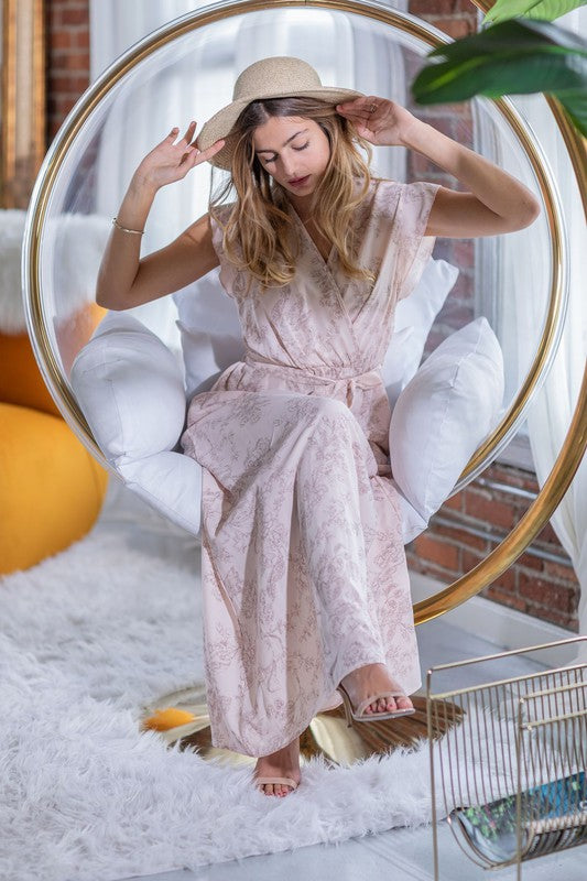 A woman in a long Floral Surplice Sash Sundress with no lining, made in the United States, stands casually in a room with contemporary furniture and large windows.