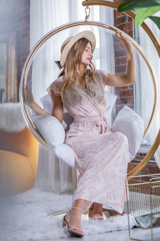 A woman in a long Floral Surplice Sash Sundress with no lining, made in the United States, stands casually in a room with contemporary furniture and large windows.