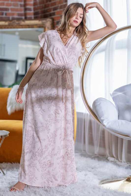 A woman in a long Floral Surplice Sash Sundress with no lining, made in the United States, stands casually in a room with contemporary furniture and large windows.