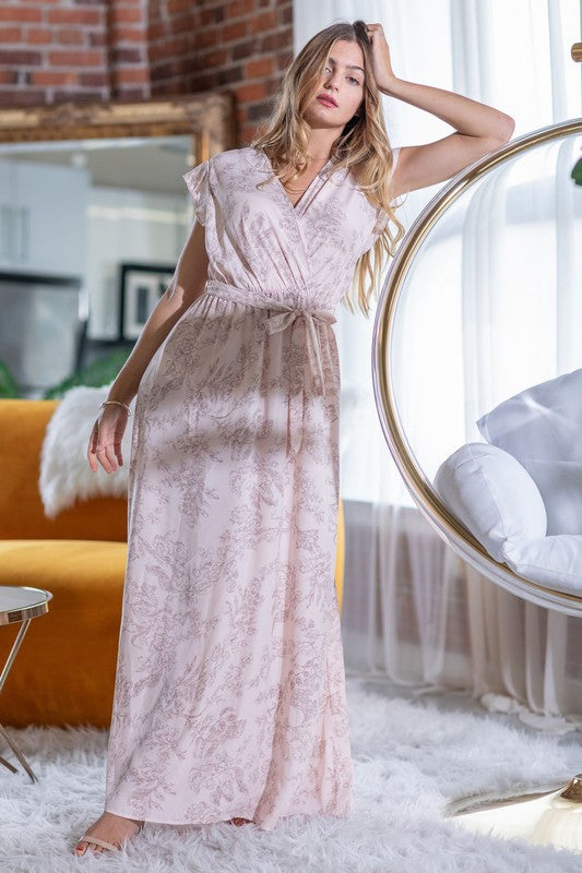 A woman in a long Floral Surplice Sash Sundress with no lining, made in the United States, stands casually in a room with contemporary furniture and large windows.