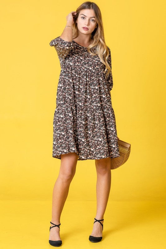 A woman in a Floral Ditsy Tiered Dress and black heels poses elegantly against a yellow background, holding a woven handbag.