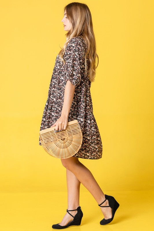 A woman in a Floral Ditsy Tiered Dress and black heels poses elegantly against a yellow background, holding a woven handbag.