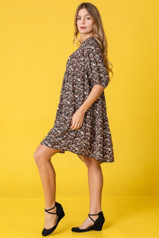 A woman in a Floral Ditsy Tiered Dress and black heels poses elegantly against a yellow background, holding a woven handbag.