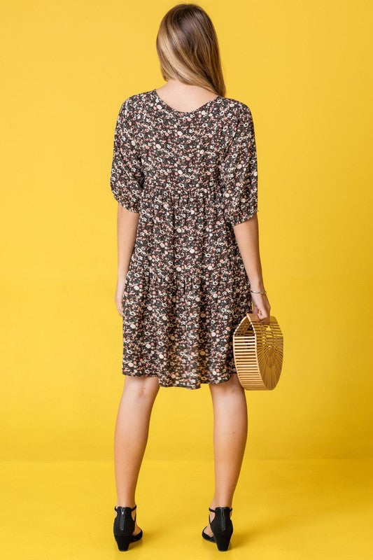 A woman in a Floral Ditsy Tiered Dress and black heels poses elegantly against a yellow background, holding a woven handbag.