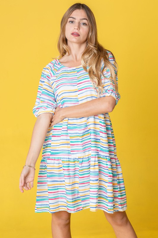 A person in a Multi Color Stripe Tiered Dress and white boots stands sideways against a yellow background. This stunning attire, proudly made in the United States, adds a pop of personality to any occasion with its playful mix of hues and styles.