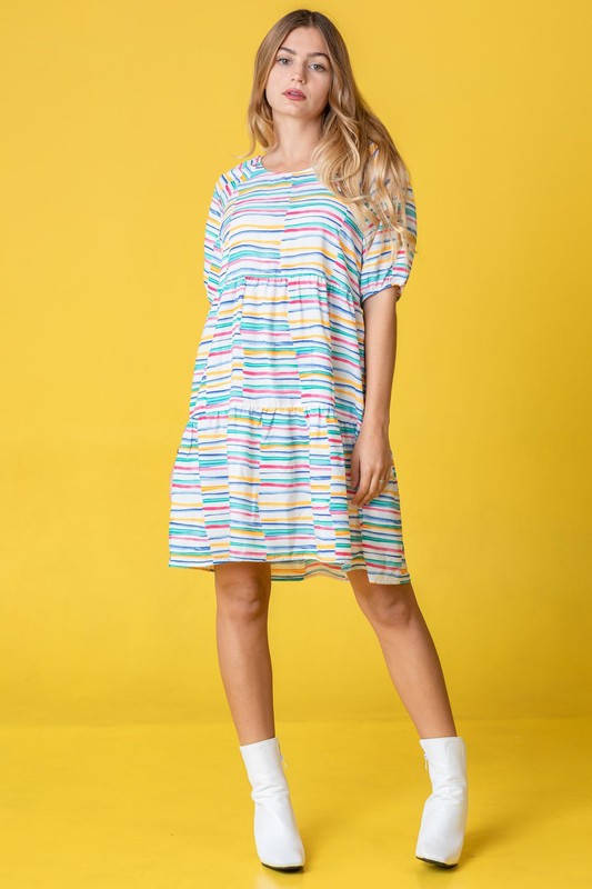 A person in a Multi Color Stripe Tiered Dress and white boots stands sideways against a yellow background. This stunning attire, proudly made in the United States, adds a pop of personality to any occasion with its playful mix of hues and styles.