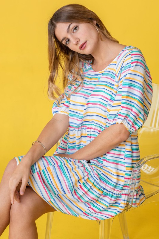 A person wearing the Multi Color Stripe Tiered Dress sits on a clear chair against a yellow background. With one hand in their hair, they look down, radiating effortless style.