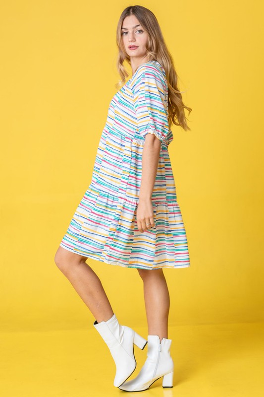 A person in a Multi Color Stripe Tiered Dress and white boots stands sideways against a yellow background. This stunning attire, proudly made in the United States, adds a pop of personality to any occasion with its playful mix of hues and styles.