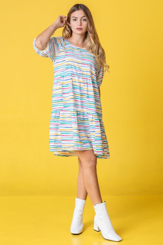 A person in a Multi Color Stripe Tiered Dress and white boots stands sideways against a yellow background. This stunning attire, proudly made in the United States, adds a pop of personality to any occasion with its playful mix of hues and styles.