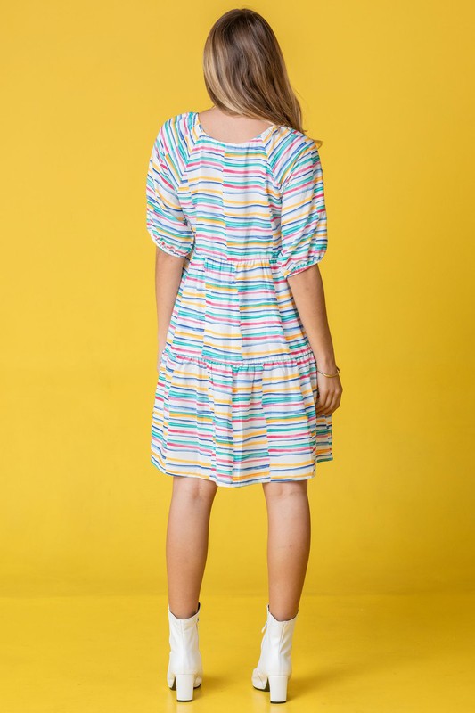 A person in a Multi Color Stripe Tiered Dress and white boots stands sideways against a yellow background. This stunning attire, proudly made in the United States, adds a pop of personality to any occasion with its playful mix of hues and styles.