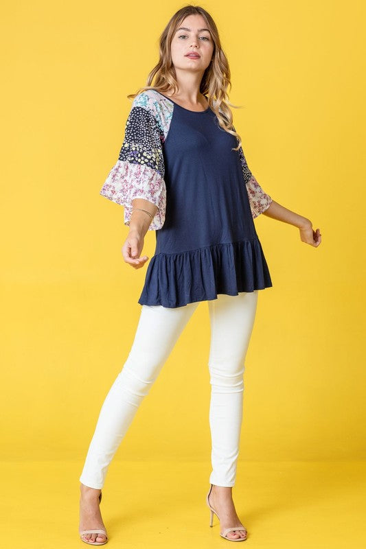 A woman stands against a yellow background wearing the Mix Pattern Circular Flounce Sleeve Tunic in navy, showcasing floral and polka dot designs on the sleeves.