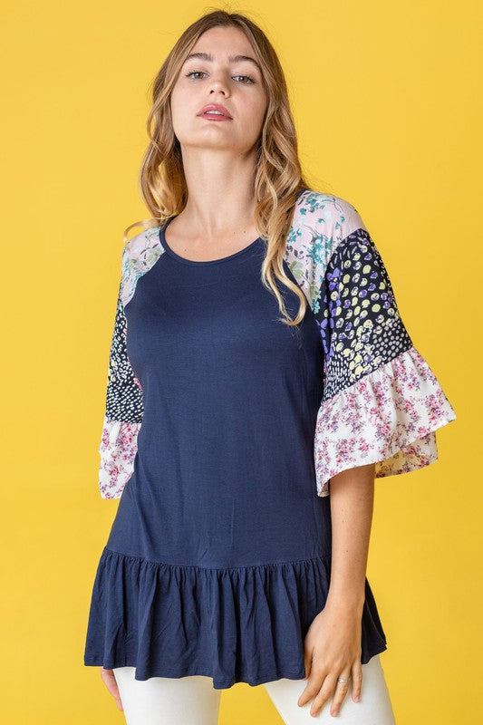 A woman stands against a yellow background wearing the Mix Pattern Circular Flounce Sleeve Tunic in navy, showcasing floral and polka dot designs on the sleeves.