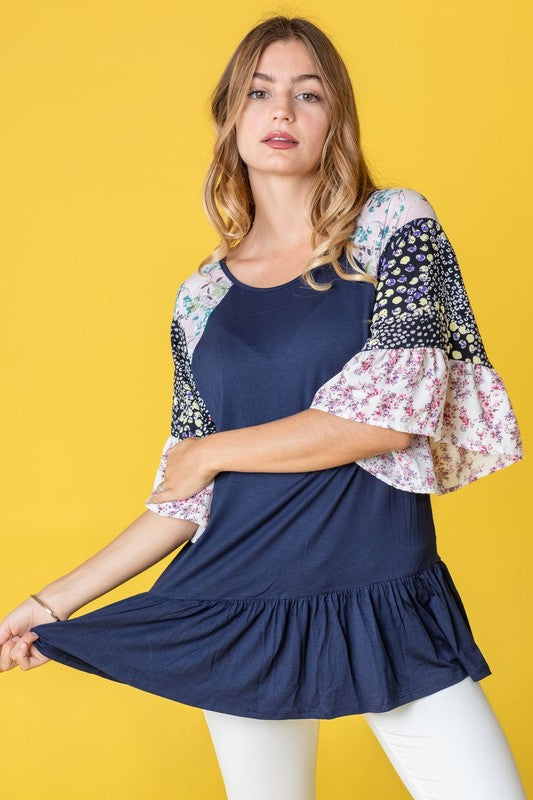 A woman stands against a yellow background wearing the Mix Pattern Circular Flounce Sleeve Tunic in navy, showcasing floral and polka dot designs on the sleeves.