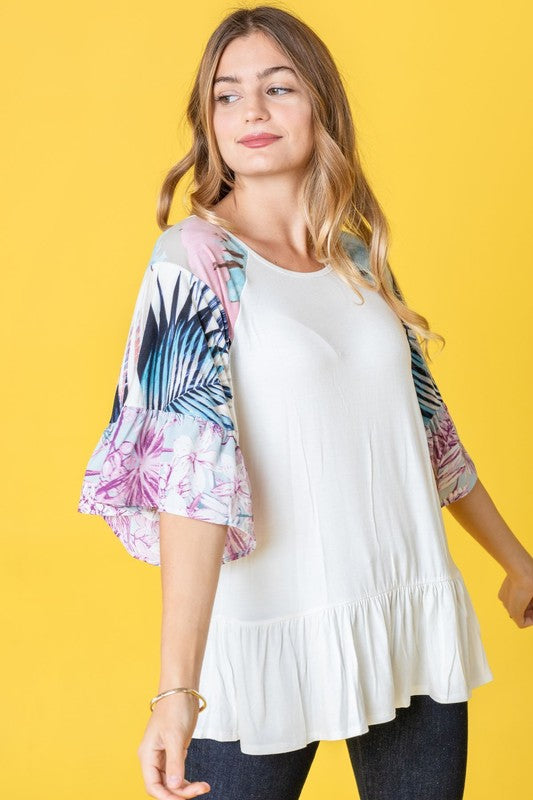 A woman stands against a yellow background wearing the Mix Pattern Circular Flounce Sleeve Tunic in navy, showcasing floral and polka dot designs on the sleeves.