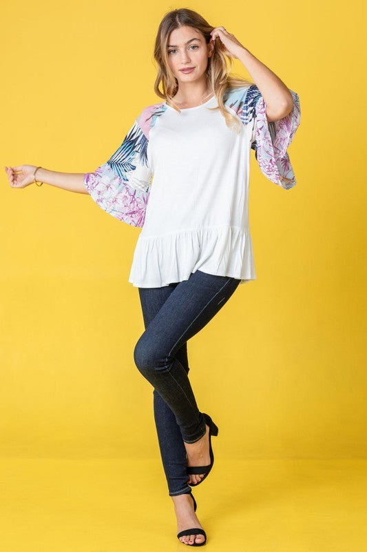 A woman stands against a yellow background wearing the Mix Pattern Circular Flounce Sleeve Tunic in navy, showcasing floral and polka dot designs on the sleeves.